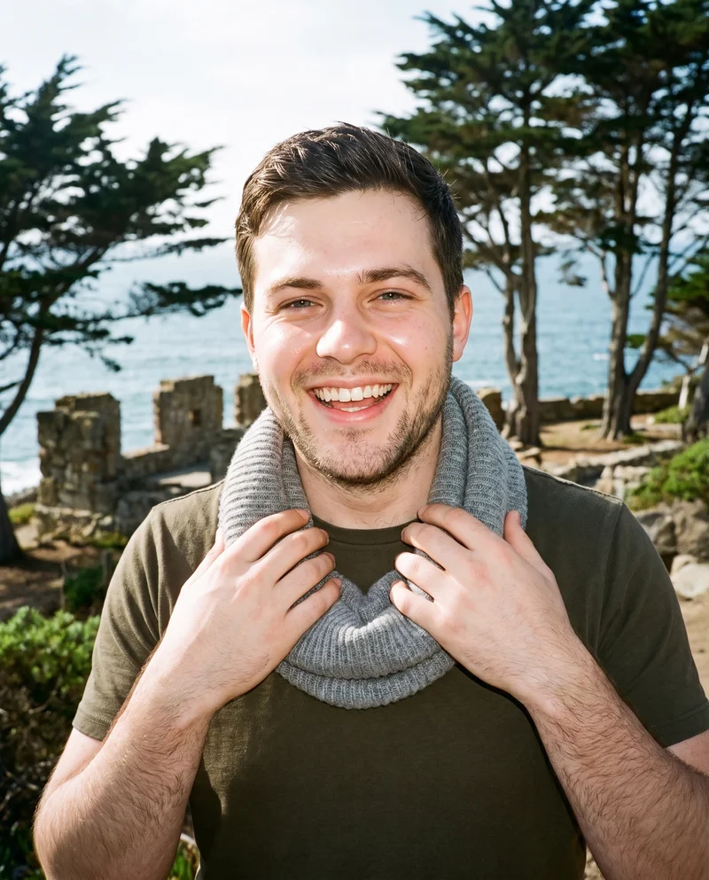 A 26-year-old Caucasian man is shown from the front in a bust frame wearing a grey knit snood, with his hands at his collarbones and a laughing expression. The Coastal Trail Cypress Ruins appear as the backdrop.