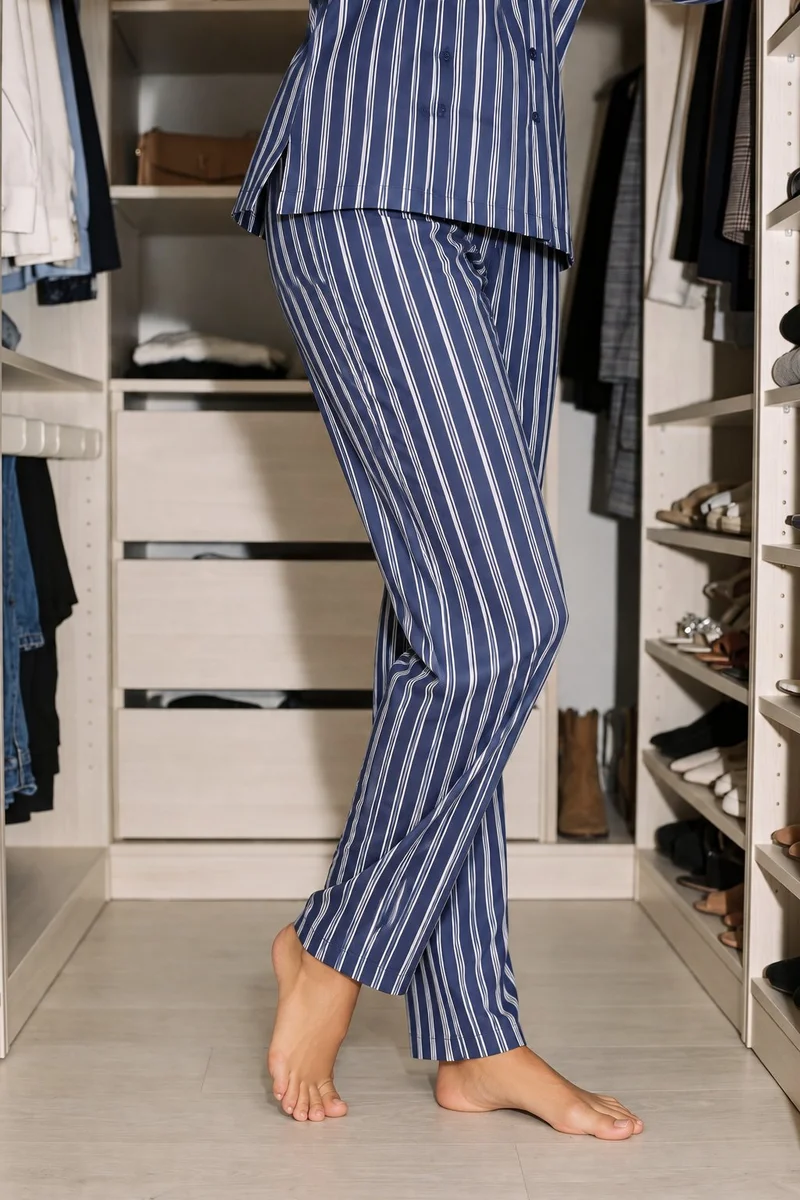 A woman wears a women's striped pyjama set in an angled front view, framed from the lower body with her ankles crossed. A walk-in closet forms the backdrop.