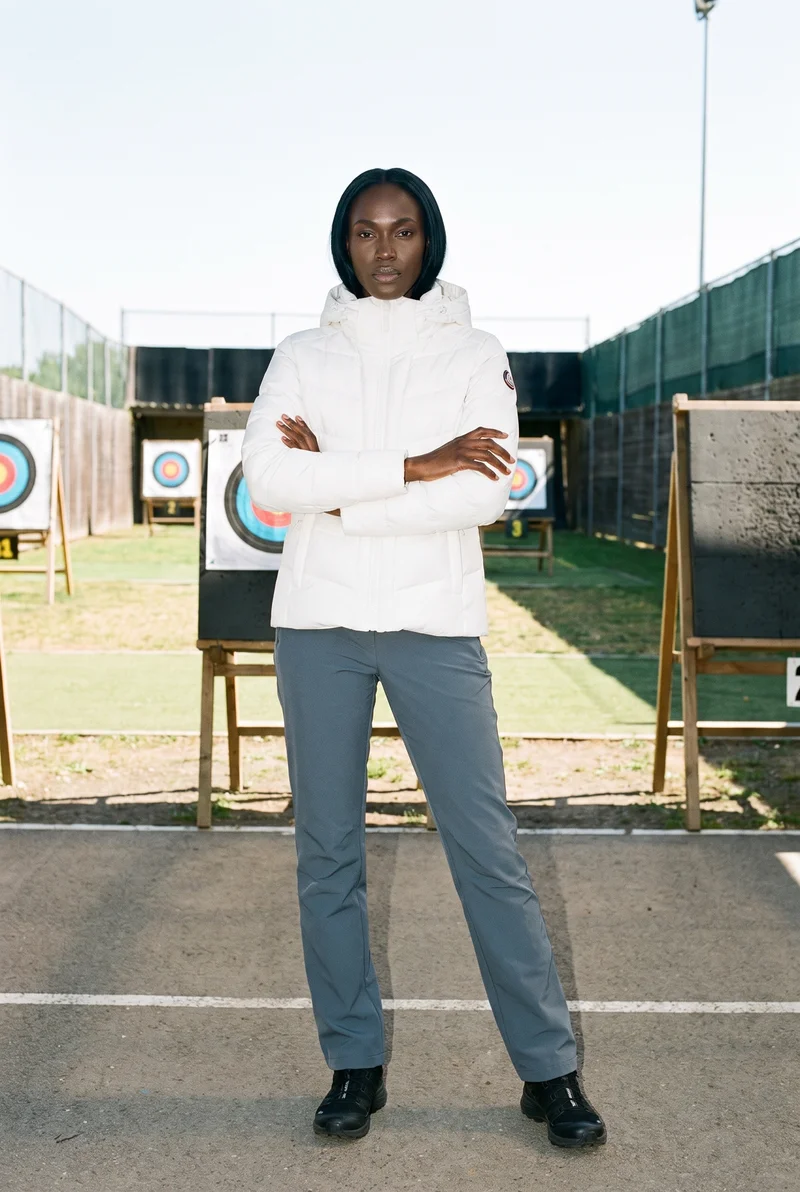 A 28-year-old African woman wears a white padded ski jacket in a full-body front view, holding herself in a self-hug pose with a neutral expression. An archery range serves as the backdrop.