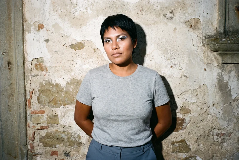 A 23-year-old Indigenous American woman is shown from the upper body in a front view, wearing a grey marl T-shirt with her hands behind her back. She has a soft facial expression, with a Tuscany hilltop village backdrop.