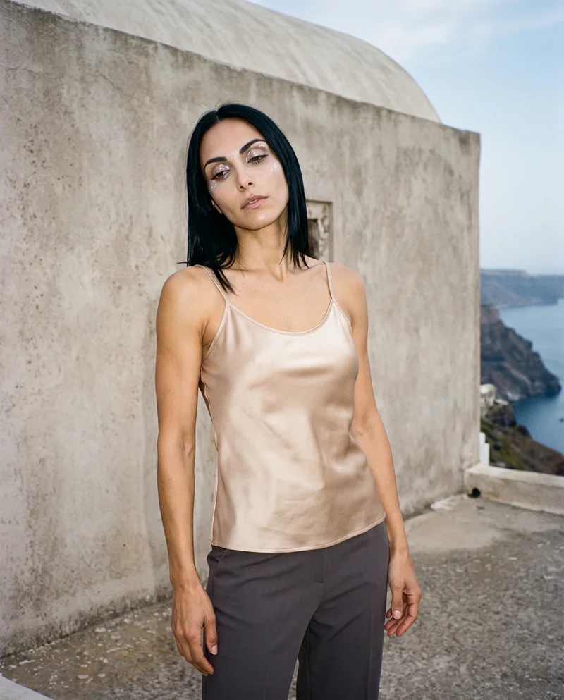 A 33-year-old Caucasian woman wears a champagne silk camisole, shown from the upper body at a front three-quarter angle with a shoulder-drop pose and a soft facial expression. A Santorini caldera view forms the backdrop.