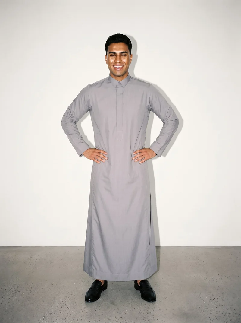 A 19-year-old Arab male model wears a grey cotton thobe in a full-body front view, standing with his hands on his hips and grinning. A soft ivory studio-wall backdrop fills the background.