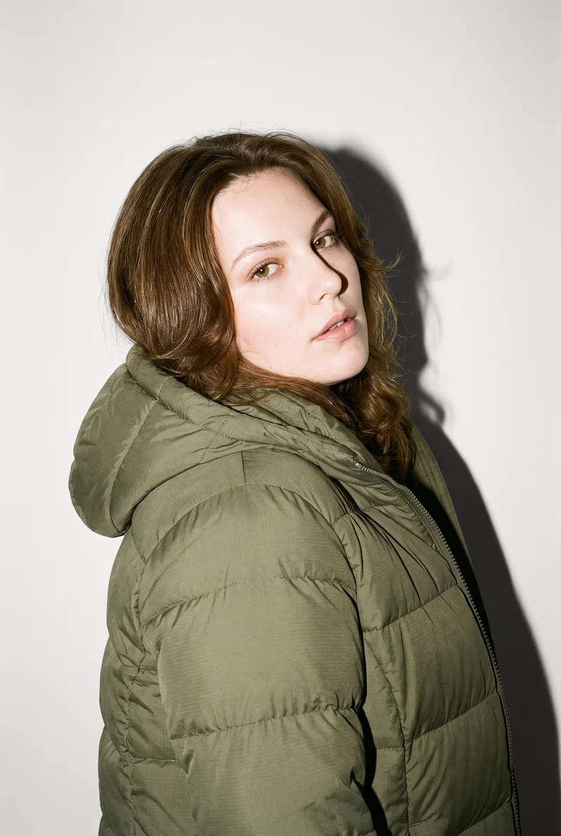 A 28-year-old Caucasian woman wears a women's olive down jacket in a bust-length front three-quarter view. She is posed with one shoulder dropped and a soft facial expression against a Chantilly Lace studio-wall backdrop.