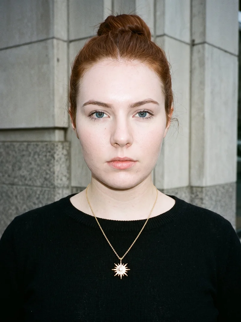 An 18-year-old Caucasian woman is shown from the bust up in a front-facing view, with her head level and a neutral expression. She wears a gold pendant necklace, set against a Financial District urban canyon backdrop.