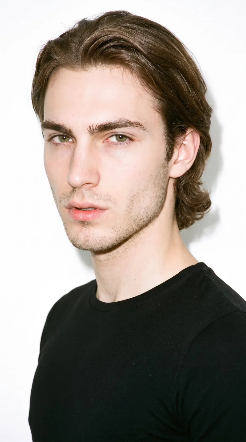 A 22-year-old Caucasian man with porcelain skin and a cool undertone has a diamond-shaped face, with a narrow forehead and jaw and cheekbones forming the widest part of the face. His eyebrows are thin, and his hazel eyes contain green-gold flecks. He has thin lips, medium-to-long neutral brown hair brushed back with movement past the ears, and very short facial-hair growth forming a faint, even shadow across the beard area.