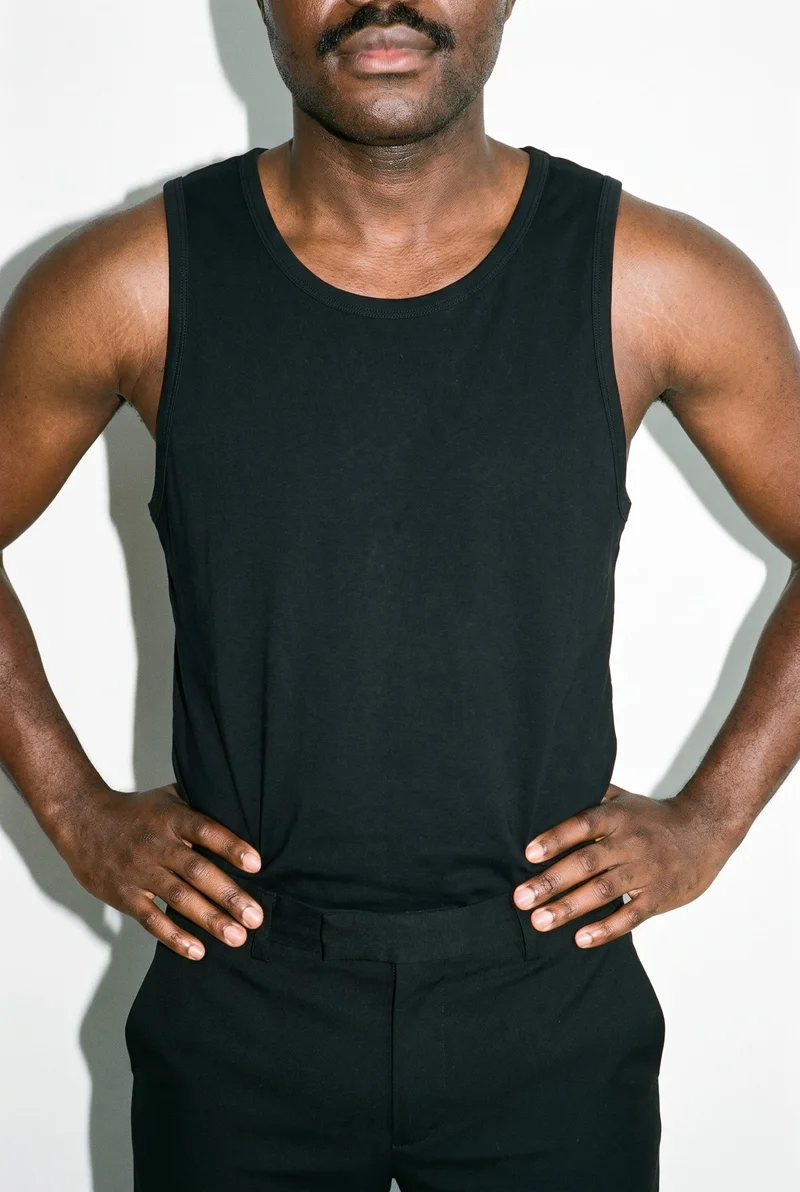 A male model is shown from the front in a torso-length view, wearing a black vest with his hands on his hips. A minimalist apartment backdrop surrounds the composition.