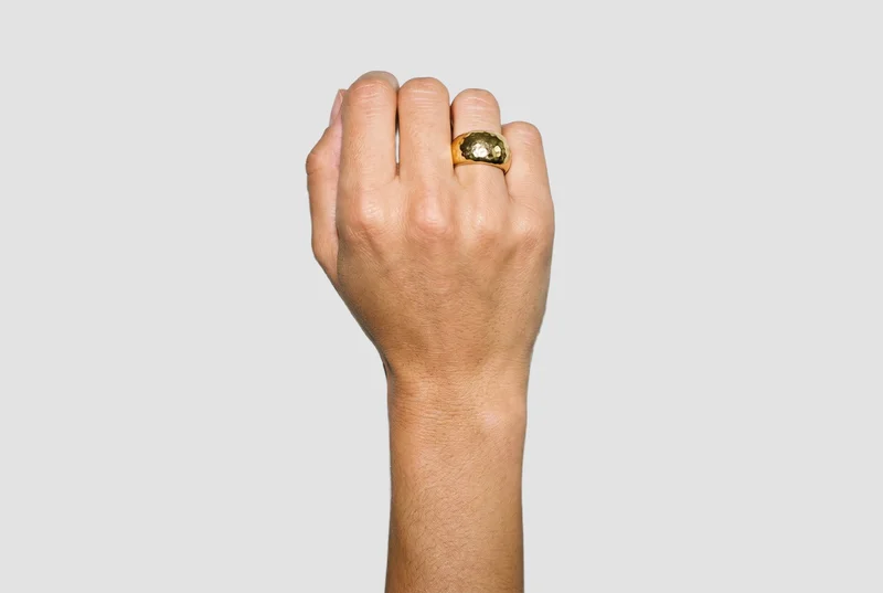 A chunky gold ring worn on a closed hand, shown with part of the wrist from a top view. The hand and wrist are framed against a flat fog-grey backdrop.
