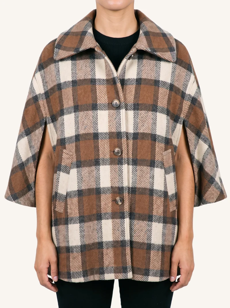 A checked wool poncho is shown from the front in a torso-length view, with the model's arms relaxed. A soft ivory flat-color backdrop surrounds the garment.