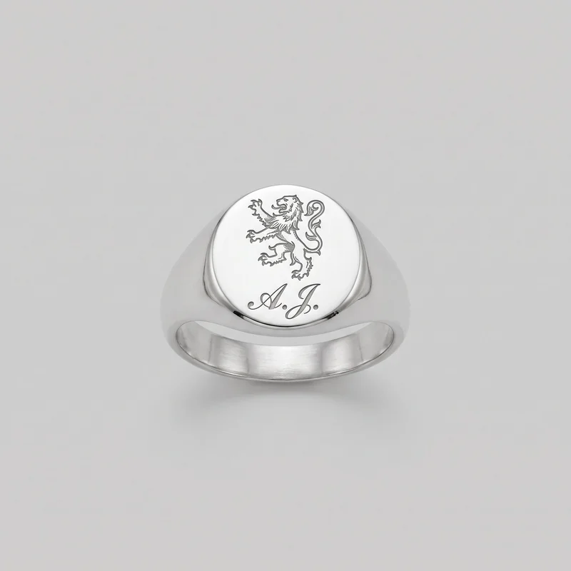A complete signet ring is shown alone from a slightly elevated front angle. Its broad tapered band widens into rounded shoulders and supports a flat circular face with a smooth raised edge. The visible exterior has rounded polished contours, an open finger hole, and a substantial underside. The face bears an engraved heraldic lion above cursive initials reading “A.J.”