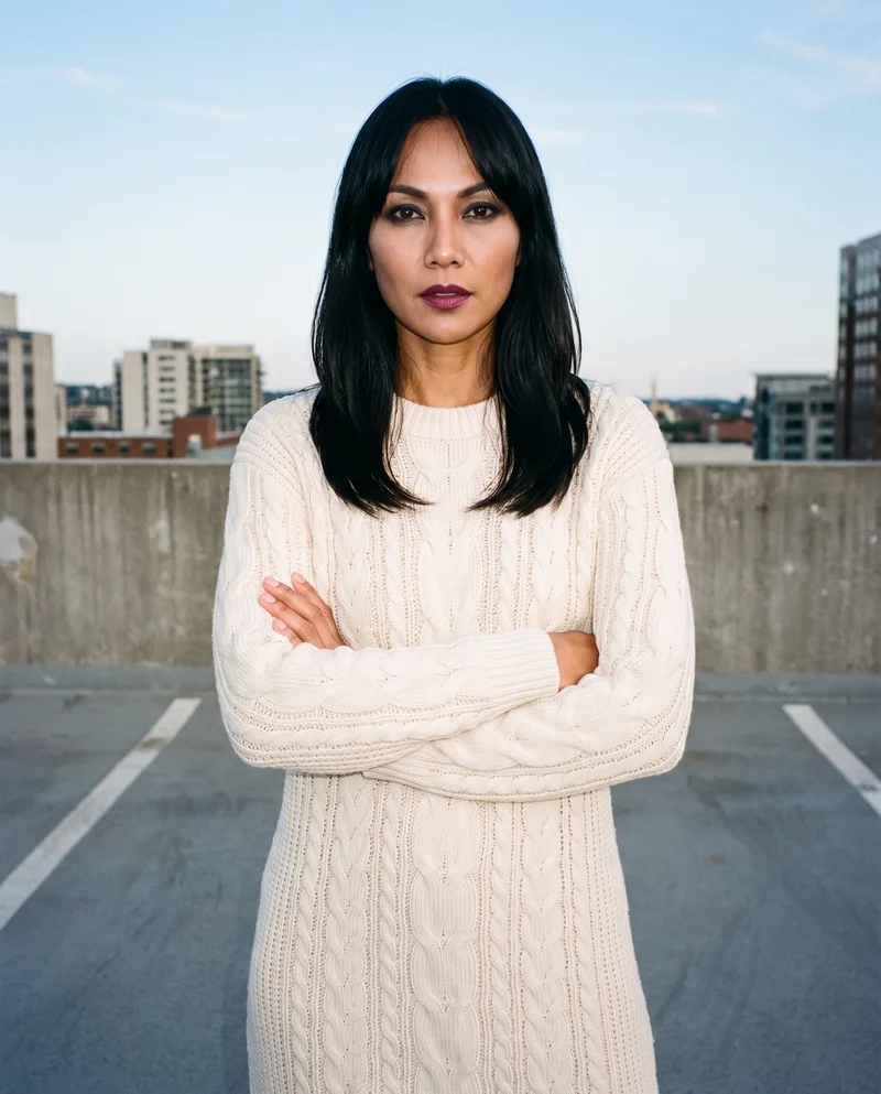 A 40-year-old Southeast Asian woman wears a cream cable-knit sweater dress in an upper-body, front-facing view with her arms crossed and a serious expression. A parking garage rooftop forms the backdrop.