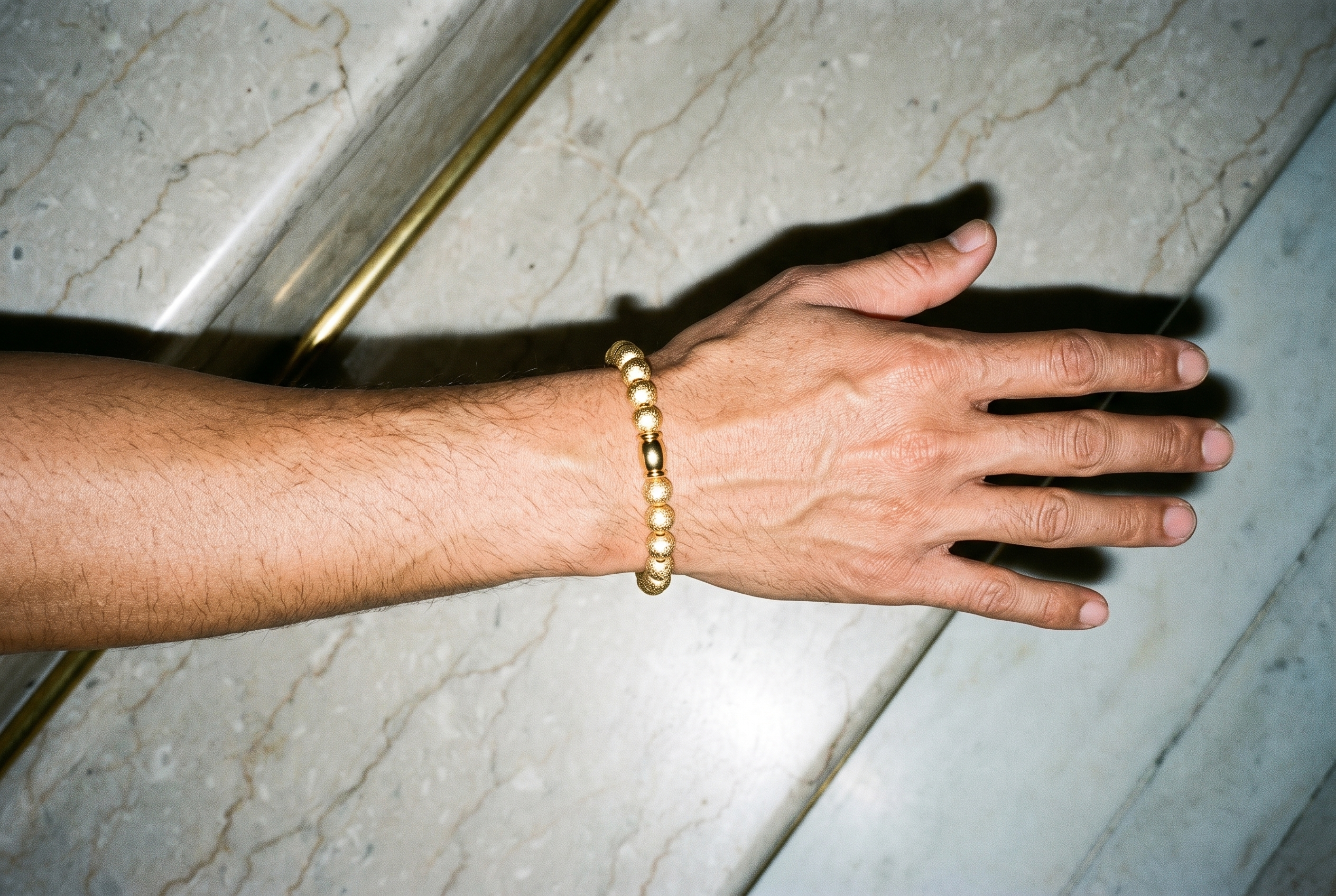 A man's hand and wrist are shown from above with the hand held flat, wearing a gold beaded bracelet. The marble staircase appears as the backdrop.
