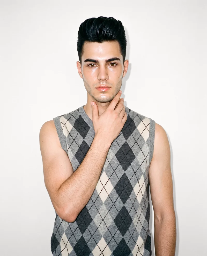 A 19-year-old Caucasian man is shown from the front in an upper-body view, wearing a grey argyle sweater vest with one hand on his neck and a natural expression. A Chantilly Lace studio-wall backdrop frames the composition.