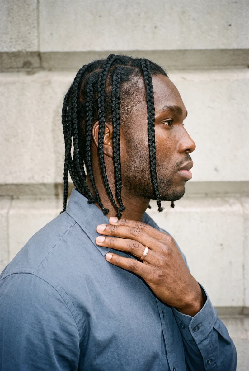 A 22-year-old African male model is shown in an upper-body side view with one hand resting at his collarbone, displaying a men's rose gold band. The natural-expression portrait uses a Financial District Urban Canyon backdrop.