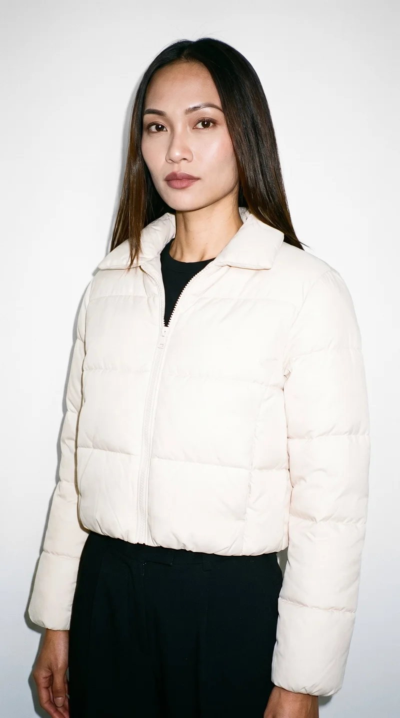 A 26-year-old Southeast Asian woman wears a cream cropped puffer jacket in an upper-body view angled from the front. Her arms are relaxed and her expression is neutral, against a pure white studio backdrop.
