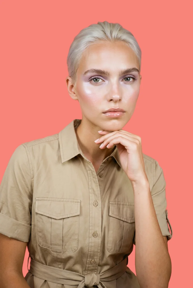 A 22-year-old Caucasian woman is shown in a bust-length front view wearing a khaki utility shirt dress. One hand rests at her jaw, and she has a serious expression against a flat coral pink backdrop.
