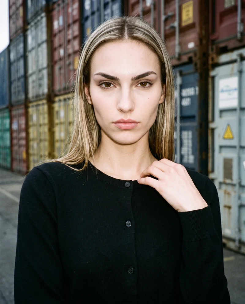 A 19-year-old Caucasian woman wears a black button-up cardigan in a bust-length front view, with one hand at her collarbone and a serious expression. Shipping containers form the backdrop.