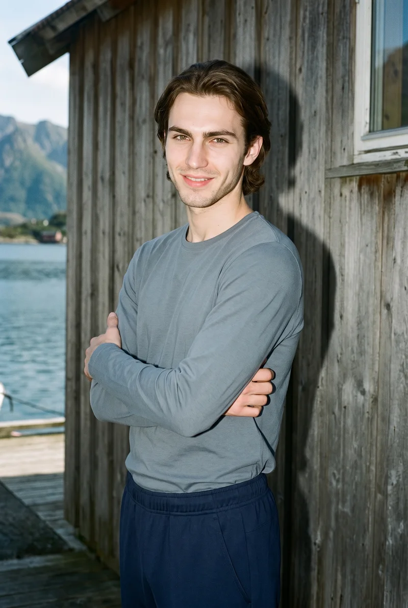 A 22-year-old Caucasian man wears navy training joggers in a three-quarter-length front three-quarter view. He is smiling and holding himself in a self-hug pose, with a Norwegian fjord village backdrop.