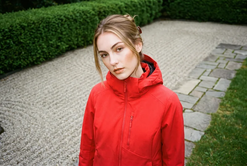 An 18-year-old Caucasian woman is shown from the upper body in a red insulated ski jacket, viewed from a front three-quarter angle. Her shoulders are lowered and she has a serious expression. A Zen rock garden serves as the backdrop.