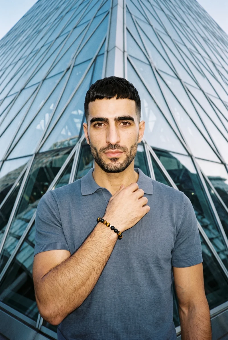 A 42-year-old Arab man is shown in an upper-body front view with one hand at his collarbone and a neutral expression. He wears a men's gemstone beaded bracelet. A pyramid building appears as the backdrop from a low-angle view.