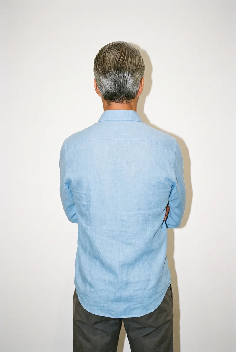 A 45-year-old Caucasian man is shown from the upper body in a back view wearing a sky blue linen shirt, with his forearms folded. A museum gallery serves as the backdrop.