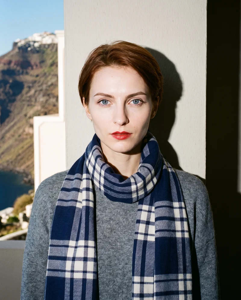 A 24-year-old Slavic woman is shown from the front in a bust composition with one shoulder forward and a neutral expression, wearing a women's navy checked wool scarf. A Santorini caldera view appears as the backdrop.