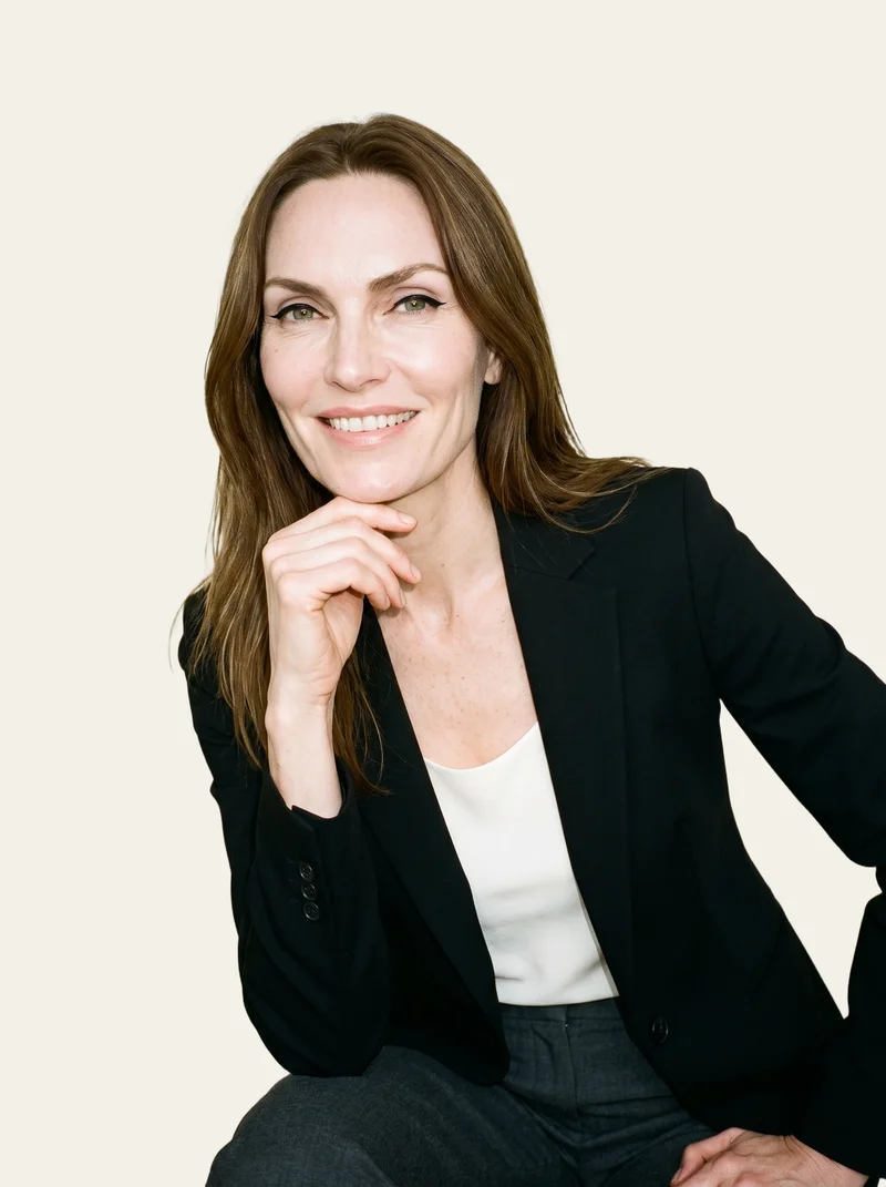 A 56-year-old Caucasian woman wears a women's black tailored blazer in an upper-body view. She is shown from a front three-quarter angle, smiling with one hand at her chin, against an alabaster flat-color backdrop.