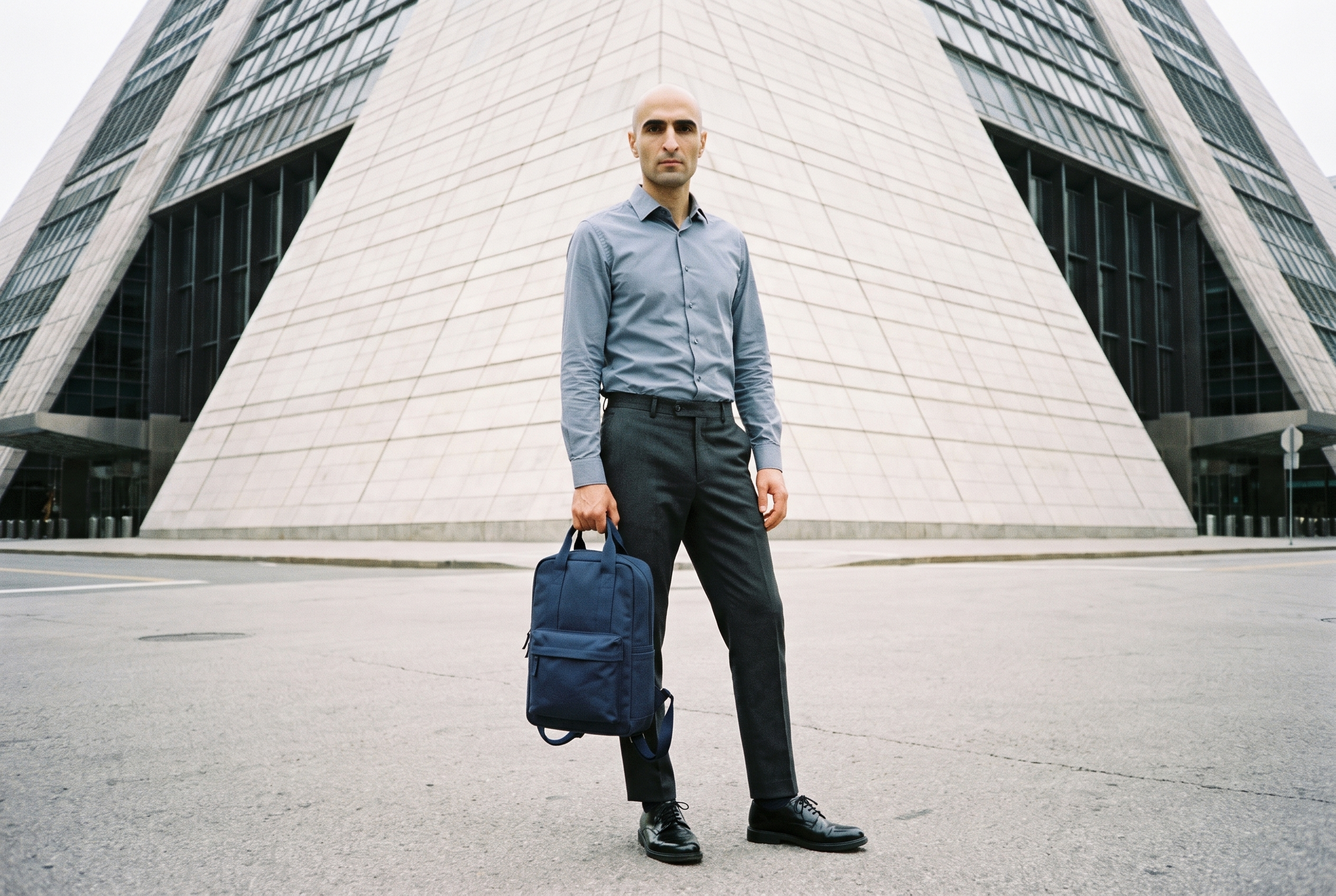A 37-year-old Slavic man is shown full body from the front with a neutral expression, displaying a men's navy canvas backpack as it dangles. A pyramid building appears as a low-angle backdrop.