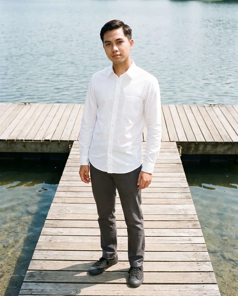 A 21-year-old Southeast Asian male model wears a white button-down shirt in a full-body front view, standing with his weight back and a natural expression. A lakeside dock forms the backdrop.