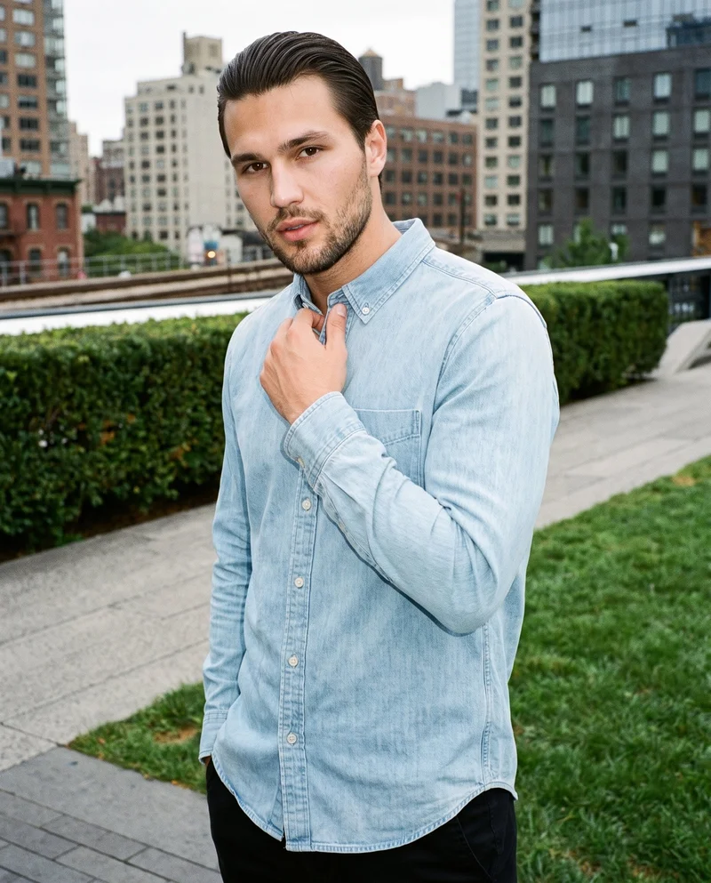 A 26-year-old Caucasian man wears a light wash denim shirt in an upper-body, angled-front view, with one hand at his collarbone and a confident expression. An elevated park railway forms the backdrop.