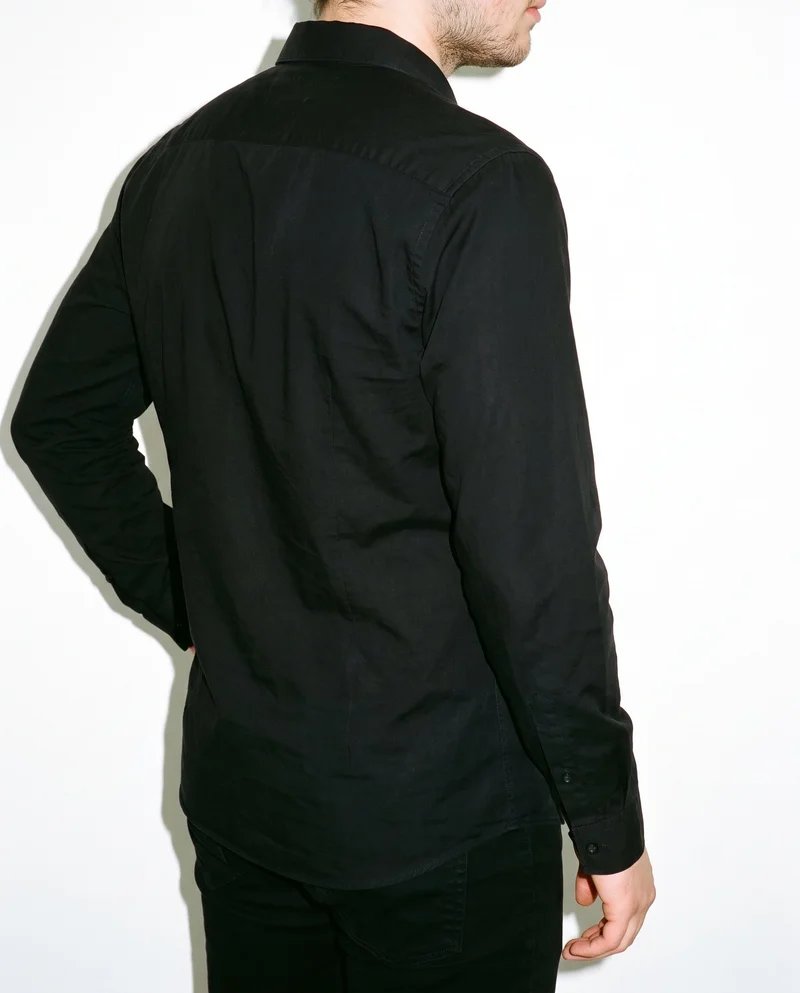 A black grandad shirt is shown on a male model in a torso-length frame, viewed from an angled rear position with one hand on his hip. A museum gallery serves as the backdrop.
