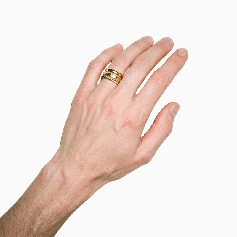 A hand and wrist are shown from above at an angle, wearing a men's gold stacking ring set. The set is presented against a flat Seasalt-colored backdrop.