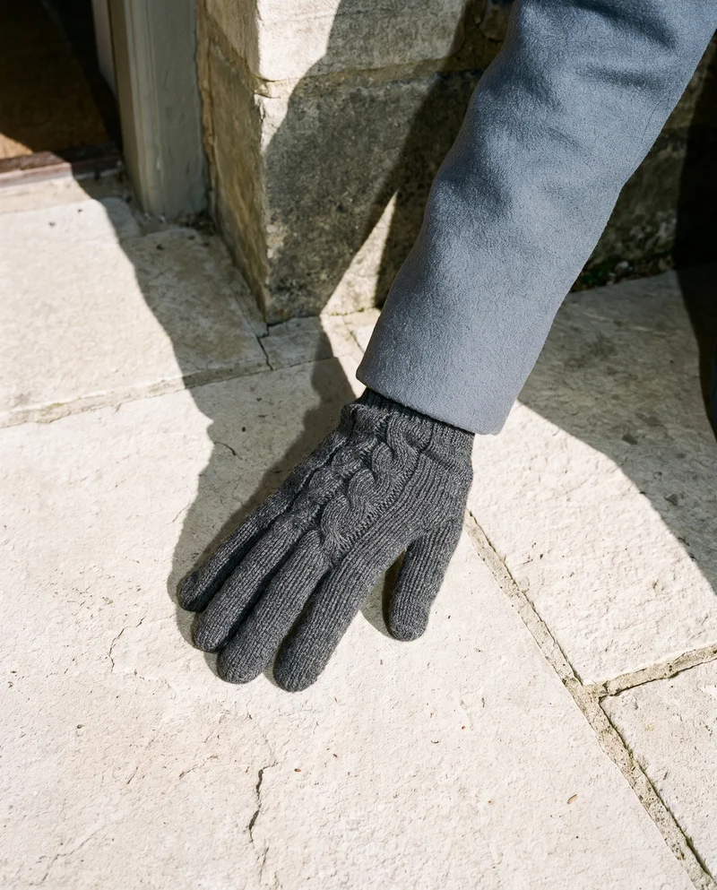 A hand and wrist wearing a grey knit glove are shown flat from above. The hand is framed against an English countryside manor backdrop.