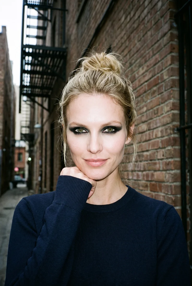 A 32-year-old Caucasian woman wears a navy merino knit in a bust-framed front view. She is smiling with one wrist raised at her jaw, against a city-alleyway backdrop.