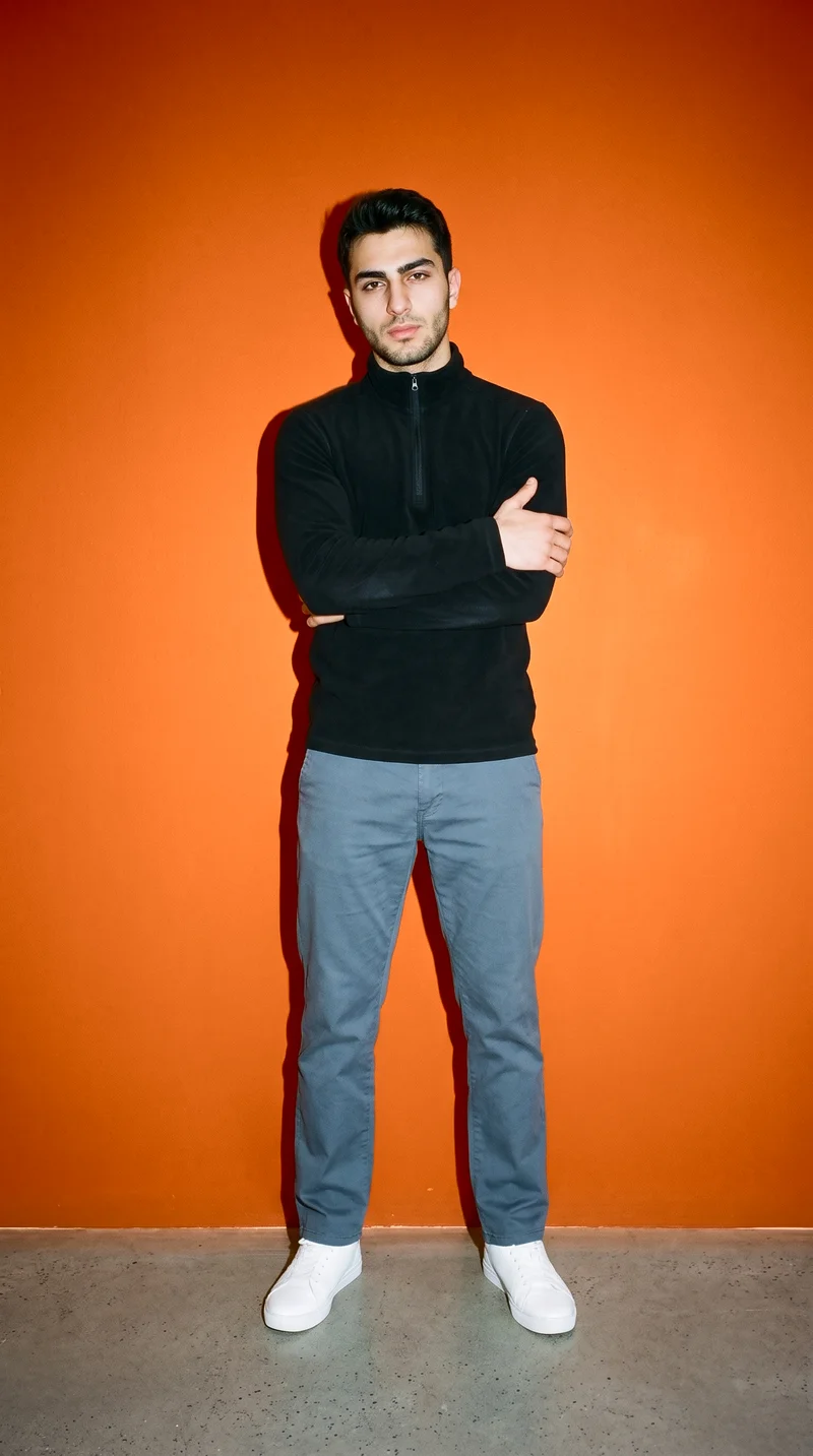 A 19-year-old Turkish man is shown full body from the front wearing a black half-zip. He stands in a self-hug pose with a natural expression against a burnt-orange studio-wall backdrop.