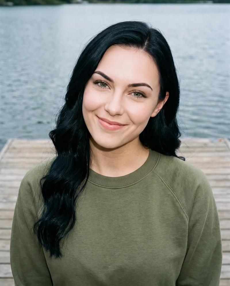 A 19-year-old Caucasian woman wears an olive crewneck sweatshirt in a bust-length front view. She is smiling with her head tilted, against a lakeside dock backdrop.