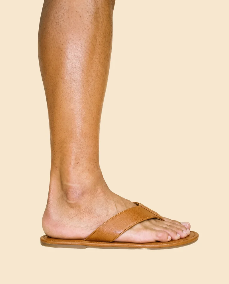 A male ankle wearing tan leather flip-flops is shown in side view with the ankle flexed. The product is framed from the ankle against a flat champagne-colored backdrop.