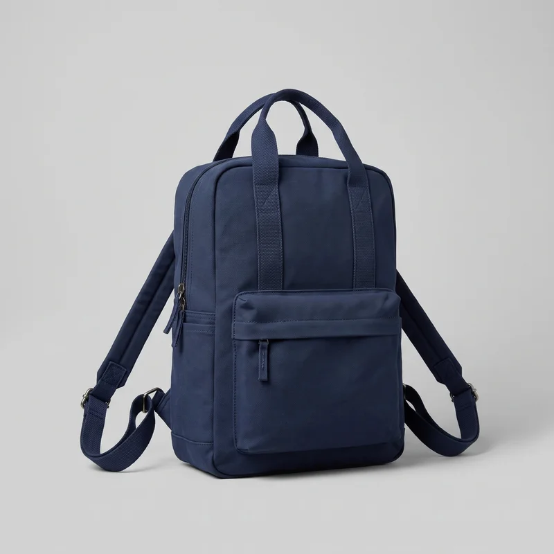 A navy canvas backpack stands upright in a front-facing view at a slight angle. Its rectangular, structured body has rounded top corners, a flat front panel, two short top handles, padded shoulder straps, a main zipper around the top and side edge, and a raised rectangular zip pocket on the lower front. Vertical webbing bands, bound edges, piping, stitched strap anchors, metal adjuster hardware, and the woven canvas texture are visible against a plain backdrop.