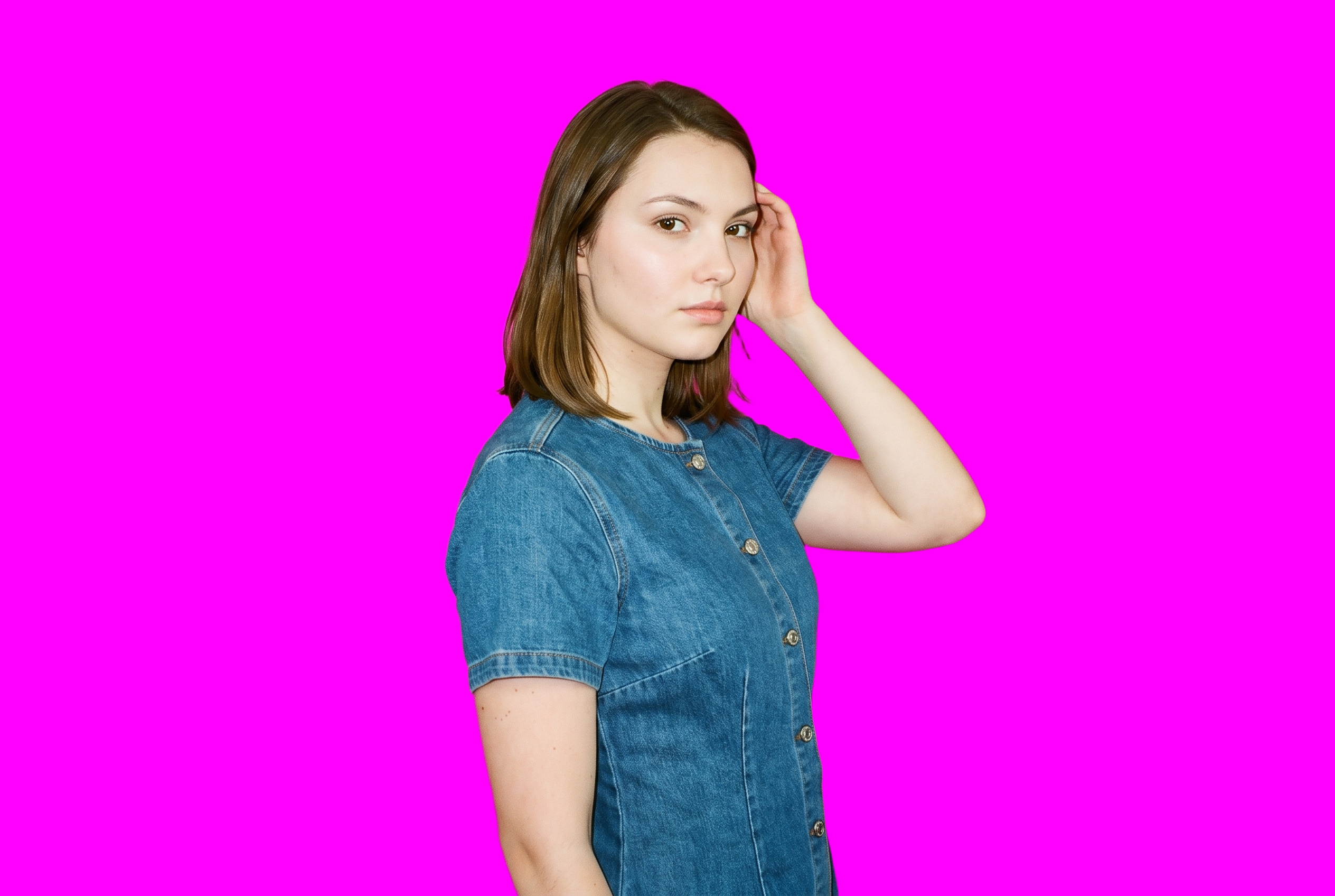 A 21-year-old Caucasian woman is shown from the upper body in a denim mini dress, viewed from a front three-quarter angle with one hand at her temple. She has a confident facial expression against a flat fuchsia backdrop.