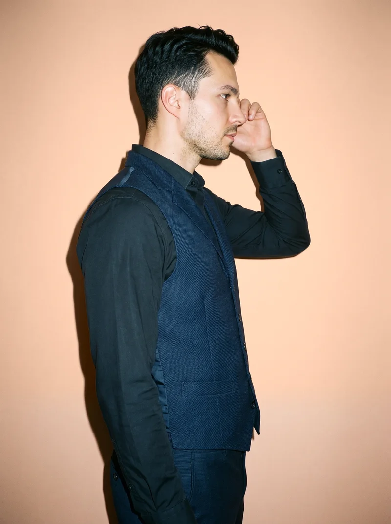 A 32-year-old Caucasian man is shown from the upper body in side view wearing a navy wool waistcoat. His hand is raised to his temple, and he has a neutral facial expression. An apricot studio-wall backdrop fills the background.