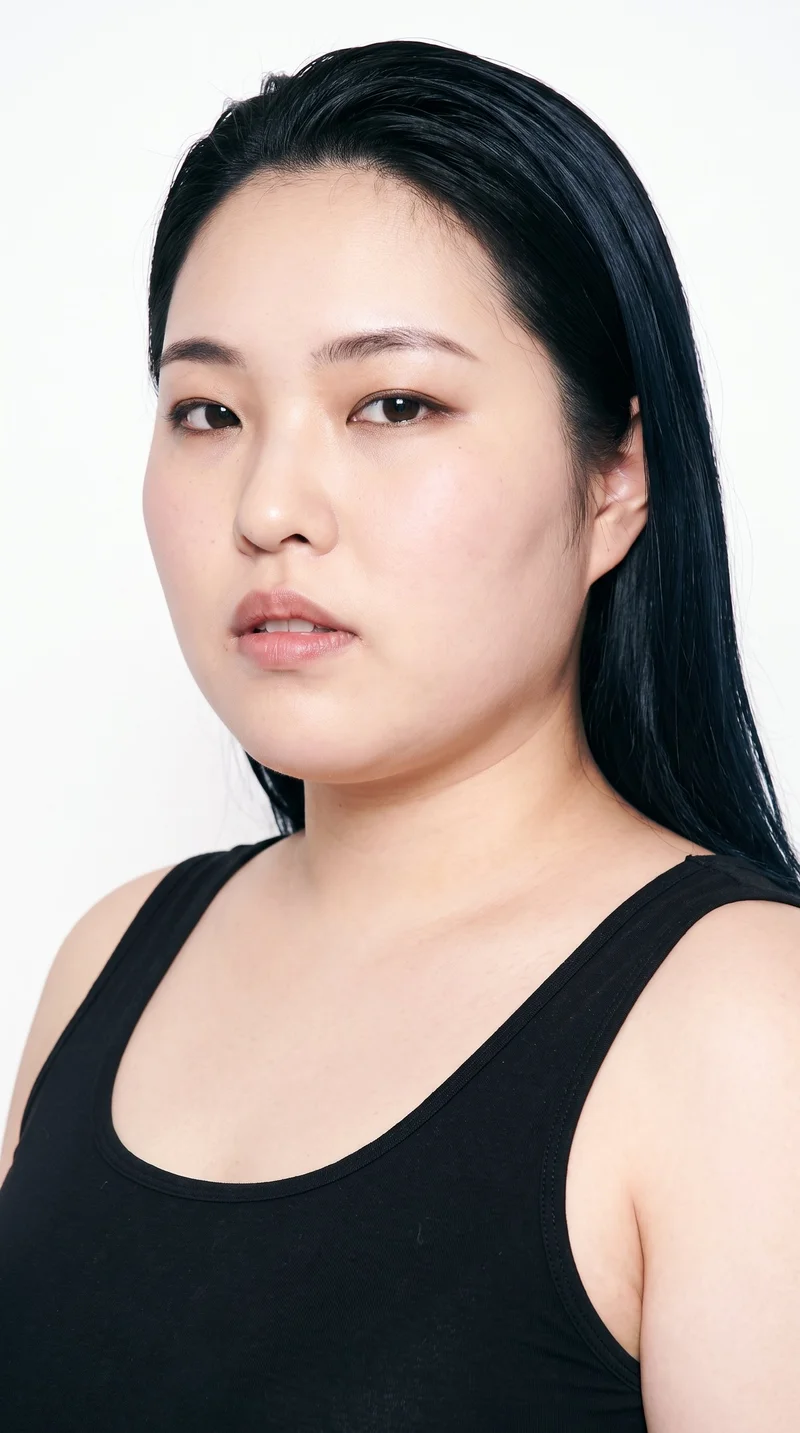 A 26-year-old Japanese woman with fair skin and a cool undertone. Her face has sharp, defined cheekbones and jawlines, creating an angular shape. She has thin eyebrows, very dark near-black eyes, and full lips. Her deep black hair is combed flat and back close to her head.