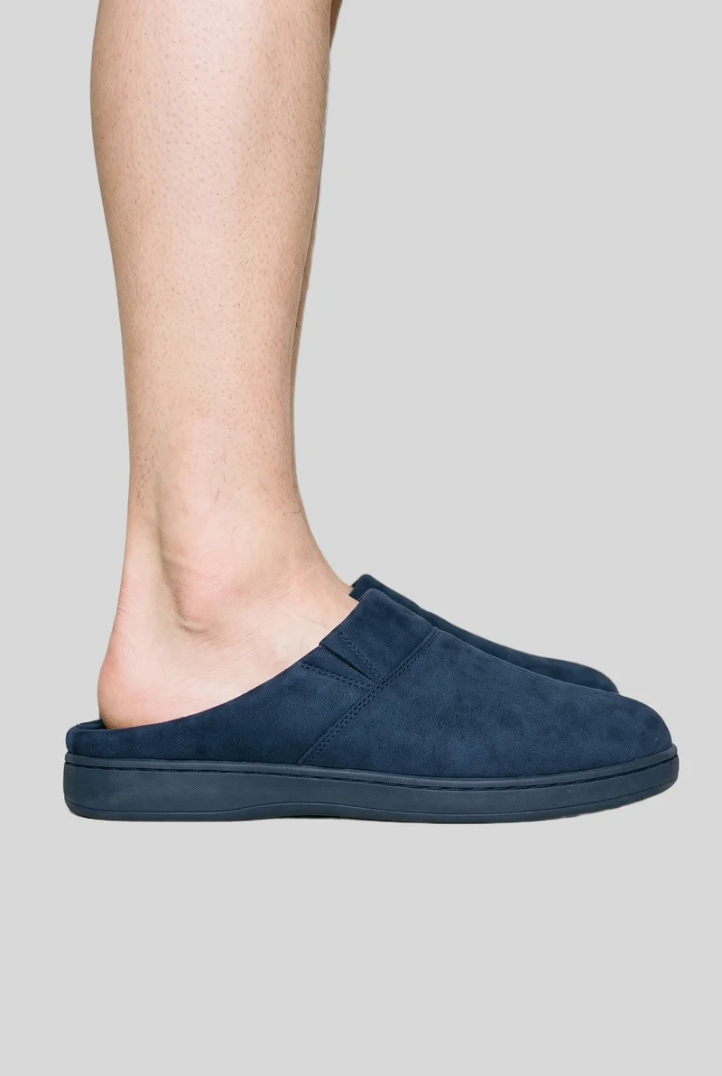 A navy mule slipper shown from the side at ankle level, framed around the ankle against a flat pearl grey backdrop.