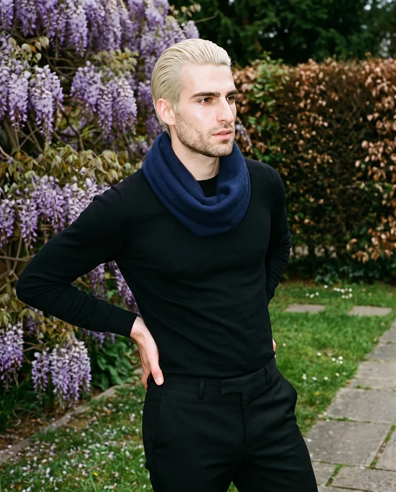 A 23-year-old Caucasian man wearing a navy snood is shown in an upper-body, angled-front view with one hand on his hip and a neutral expression. A wisteria archway forms the backdrop.