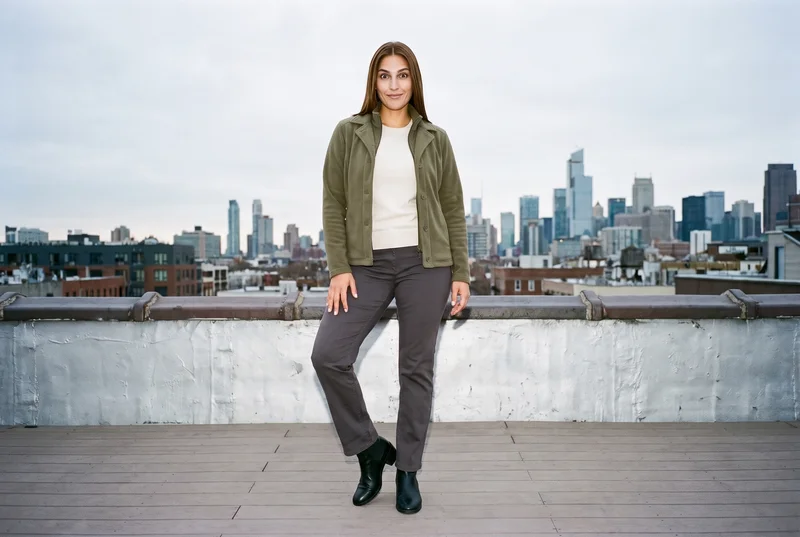 A 31-year-old Hispanic woman models an olive fleece jacket in a front-facing environmental view, standing with one hip out and a curious expression. A warehouse rooftop skyline serves as the backdrop.