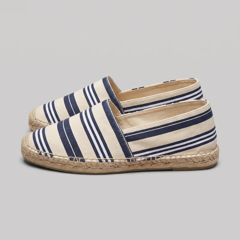 A pair of men's striped espadrille flat shoes is shown from an angled side view, with one shoe mostly visible and the second positioned behind it. The slip-on shoes have horizontal stripes, an open topline, rounded toes, and braided espadrille-style edges around the flat soles.