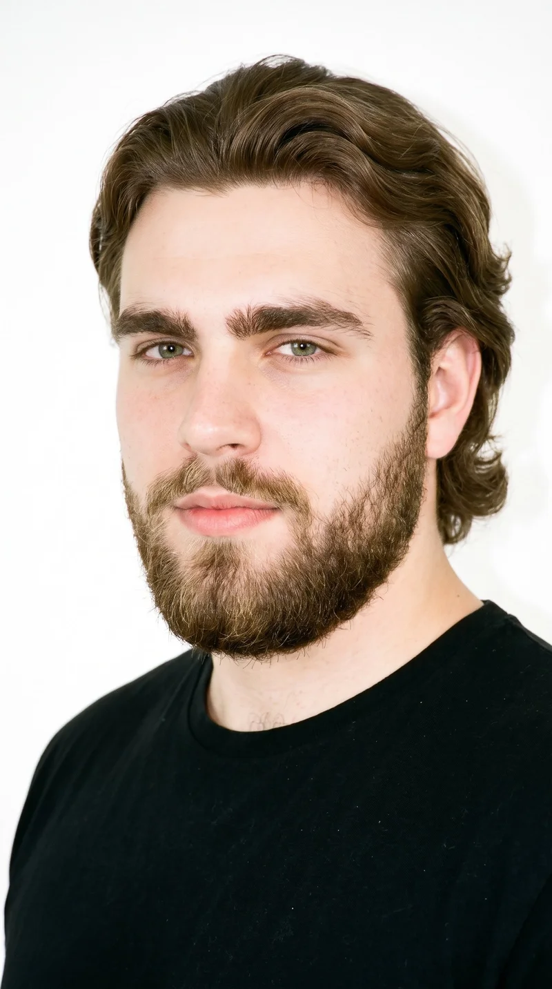 A 19-year-old Slavic man with porcelain skin and a neutral undertone has a square, angular face with a broad forehead and strong, defined jaw. His thick eyebrows frame green eyes, and his medium lips sit above a full beard grown evenly and continuously across the cheeks, jaw, and chin. Brown hair of medium to long length is brushed back, with body and movement extending past the ears.