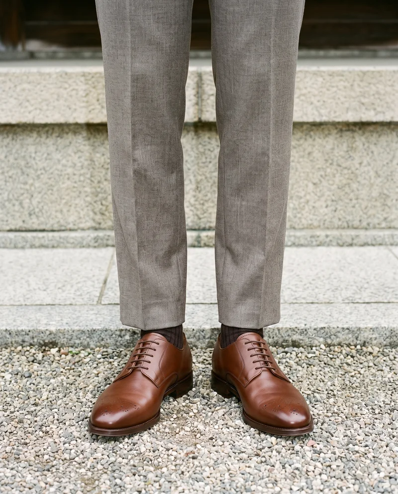 Brown derby shoes worn by a male model, shown from the front with the feet together in a lower-body view. A Kyoto temple garden forms the backdrop.