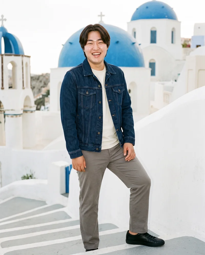 A 20-year-old Korean man wears a men's indigo denim jacket in a full-body view from a front three-quarter angle. He is laughing and standing with his weight back, against a backdrop of white Santorini steps.