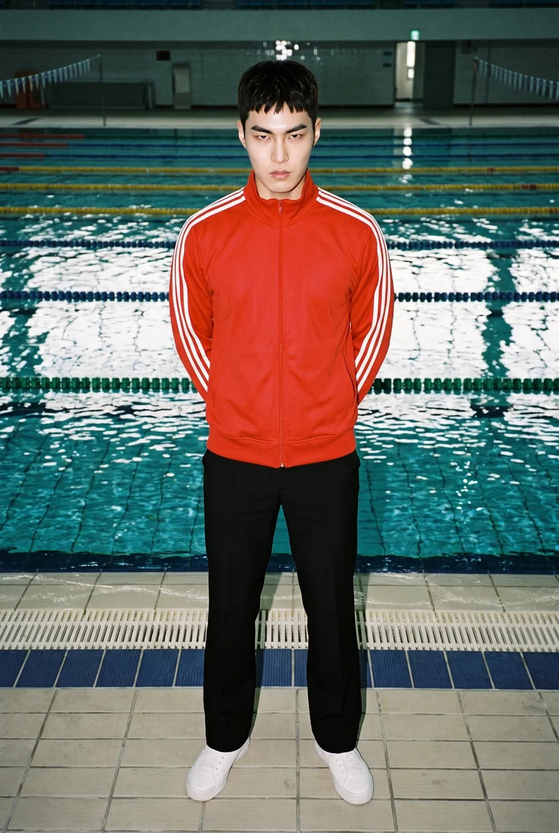A 26-year-old Korean man is shown full body from the front wearing a red striped track jacket, with his hands behind his back and an intense expression. A swimming pool Olympic backdrop is visible behind him.