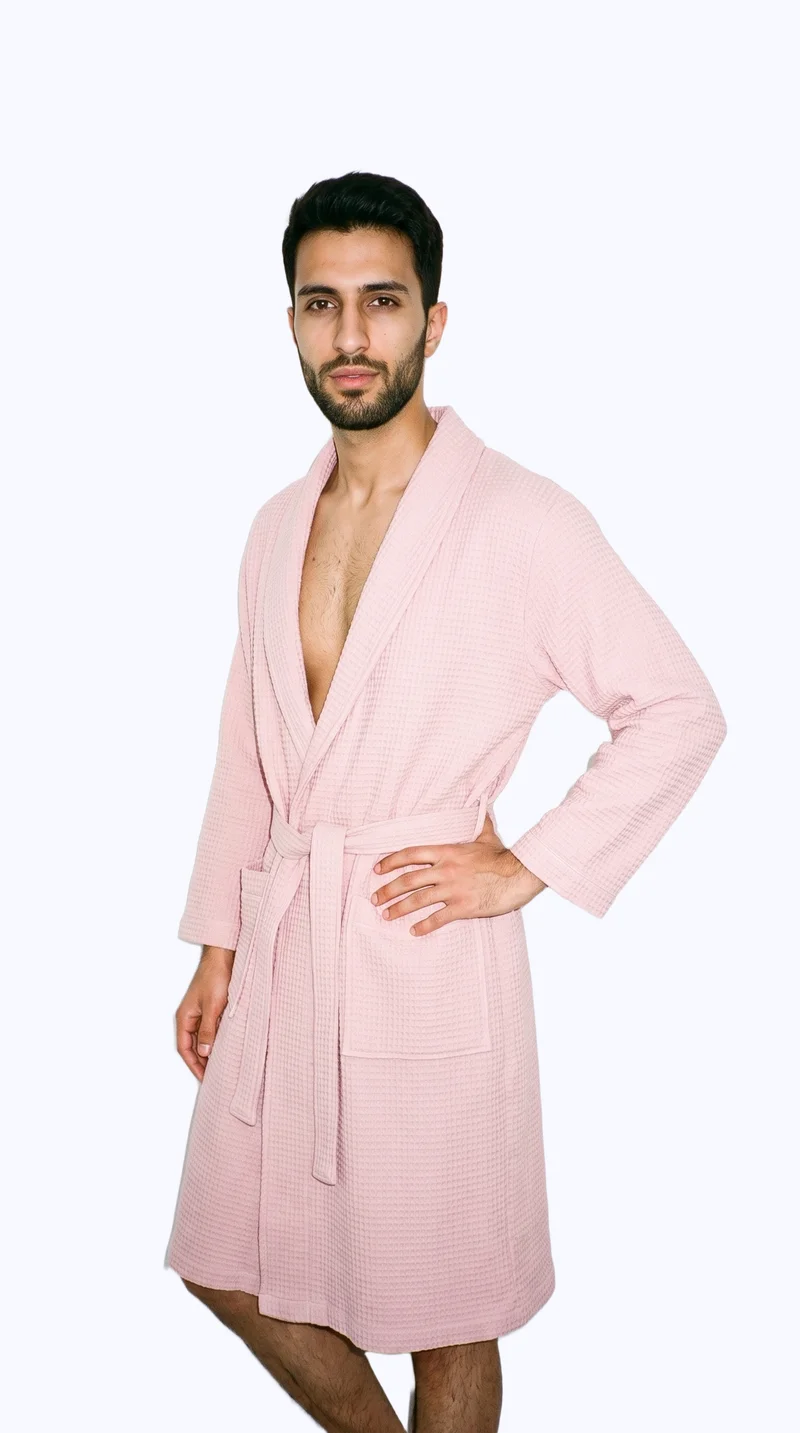 A 24-year-old Arab man wears a blush pink robe in a three-quarter-length composition. He is shown from a front three-quarter angle with his weight shifted back and a natural facial expression against a flat porcelain-color backdrop.
