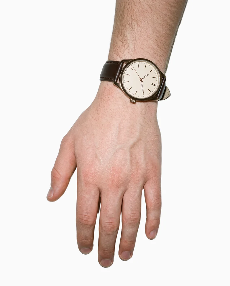 A man's wrist and hand are shown from above, with the fingers draped and a brown leather analogue watch worn on the wrist. The image is framed tightly around the hand and wrist against a flat seasalt-colored backdrop.